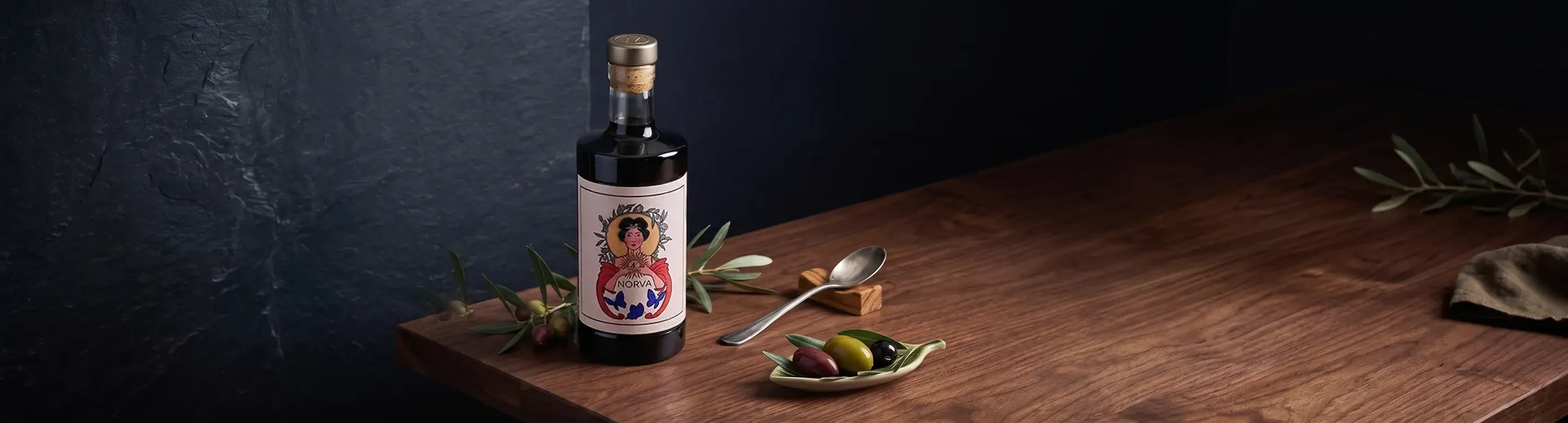 Botella de aceite de oliva virgen extra NORVA sobre una mesa de madera, con ramas de olivo, una cuchara y un plato de aceitunas.